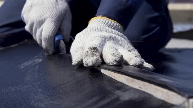 Close up for worker hands in white protective gloves cutting a black sheet of ruberoid with a technical cutter. Clip. Worker cutting out a piece from a roll of roofing material.
