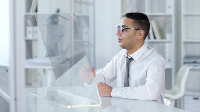 Chest-up shot of young Middle Eastern man in white shirt, tie and glasses, sitting in office and browsing through information on transparent VR touchscreen with swiping gestures. Suitable for AR, VR.