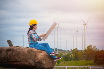 A female engineer with yellow helmet sitting and read the blueprint, Smile engineer women with laptop and wind turbine background, Happy work and planning for systematic work concept.