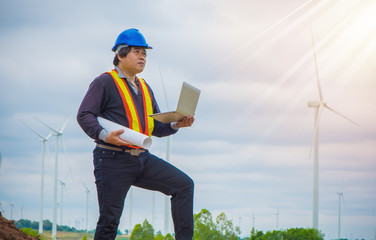 Engineer with blue helmet holding laptop and blueprint, Very happy and confident man, Look forward Smile engineer with wind turbine and sunshine background, Leadership and Successful concept.