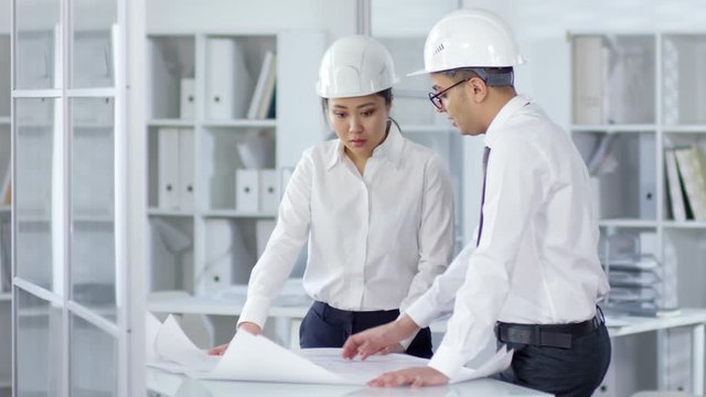 Medium shot of Asian female and Middle Eastern male engineers in business attire and white hardhats standing in office, studying technical drawings on large paper sheets and discussing details.