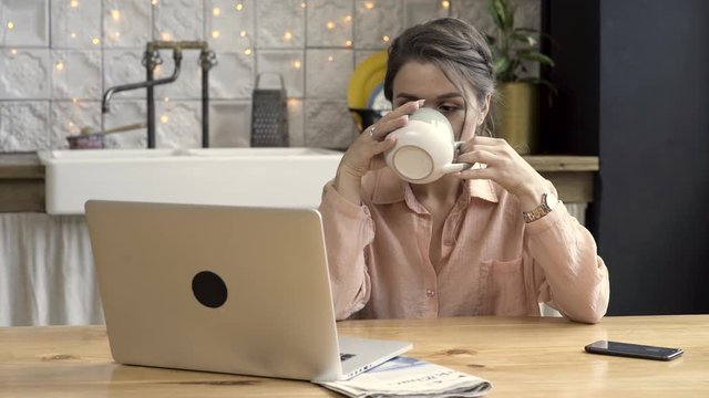 Woman working from home using laptop In the kitchen. Stock footage. Cute brunette woman drinking tea from grey and beige mug, working on laptop, freelance concept.