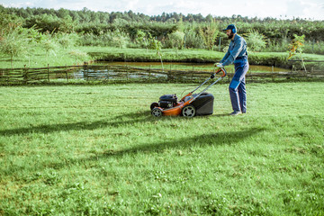 Professional gardener in protective workwear cutting grass with gasoline lawn mower on the beautiful spacious backyard