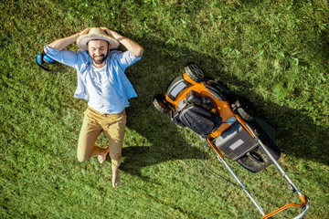 Handsome man resting on the grass near the lawn mower enjoying gardening on the backyard in the...