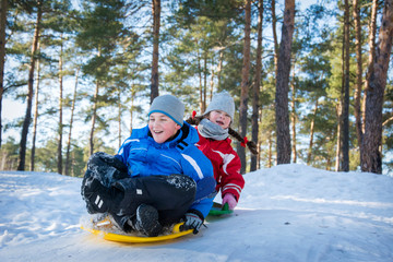 In winter, in the forest on a bright sunny day, brother and sister ride plastic plates from the mountain.