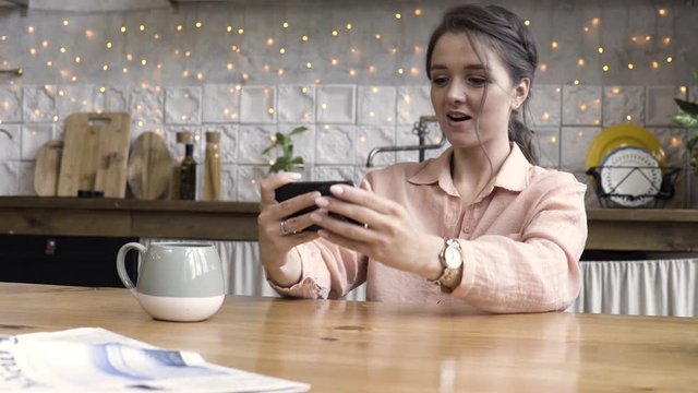 A portrait of a happy young brunette woman wearing light pink shirt and golden watch playing games on mobile phone at her kitchen. Stock footage. Modern gadget in female hands.