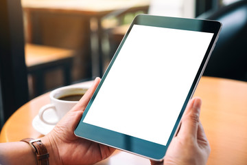 Mockup image of hands holding black tablet pc with blank white screen with coffee cup on wooden table