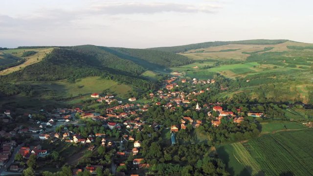 An aerial drone shot over a village in the Romanian countryside. The village of red roofs is dotted around the flat land surrounded by green fields and hills, the sun sets on the scene.
