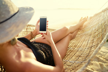 Vacation and technology. Close up of woman in hat and swimsuit using smartphone laying in hammock on the beach. Focus on screen.