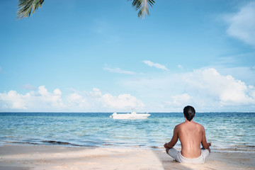 Suntan and meditation. Enjoying vacation.. Relaxed young man siiting on lotus position on the tropical beach.