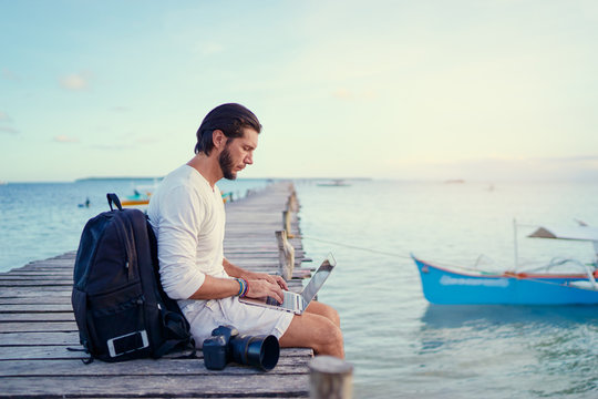 Work and travel. Young man with rucksack using laptop computer sitting on wooden fishing pier with beautiful tropical sea view.