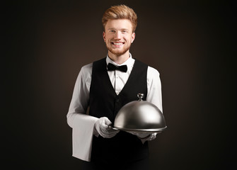 Handsome waiter with tray and cloche on dark background