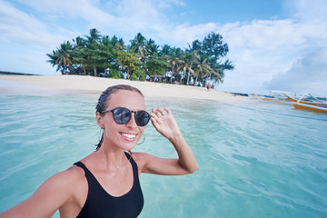Traveling in Philippines. Happy young woman taking selfie while bathing near small island with white sand tropical beach.