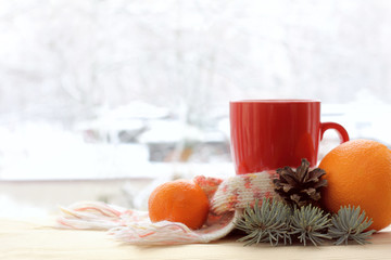 mug with a scarf, spruce branch, pine cone, orange, mandarin on a table by the window with a winter landscape. Christmas still life