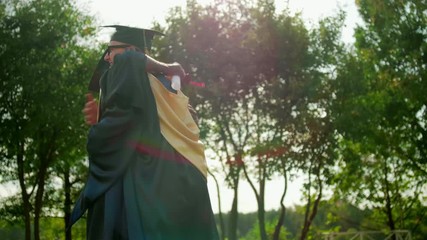 Two (African and Caucasian) Happy Graduate Students in Gowns and Caps Hug Outdoors after Diploma Graduation Ceremony. 4K Slow Motion Medium Shot with Beautiful Sun Lens Flare