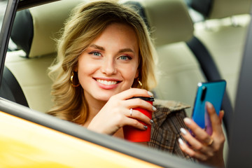 Photo of woman with coffee and phone in hands sitting in back seat of yellow taxi in summer on...