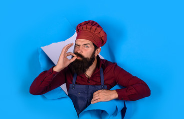 Bearded chef in uniform. Cook with taste approval gesture through paper. Chef, cook, baker gesturing excellent. Chef in hat and apron with perfect sign. Professional chef man shows sign for delicious.