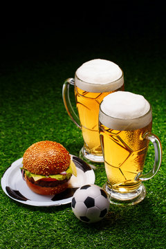 Two glasses of beer, hamburger and soccer ball on green grass.