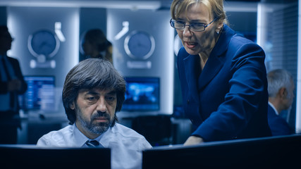 Female Manager Talks with Professional Operator Working on a Personal Computer in the System Monitoring Room. High Profile Specialist Working.