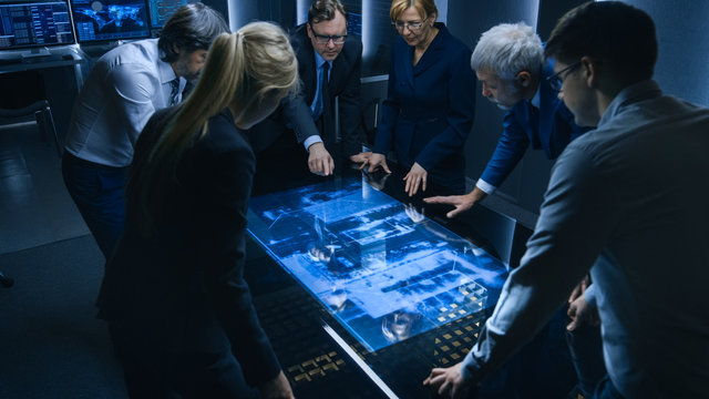 Team of Government Intelligence / FBI Agents Standing Around Digital Touch Screen Table and Tracking Suspect Vehicle Using Satellite Surveillance in the Monitoring Room.