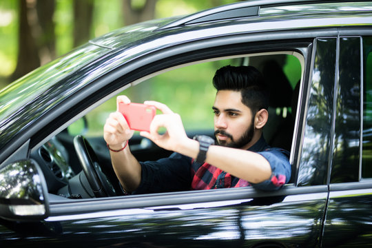 Indian driver taking photo with camera smartphone driving in car. Happy man taking picture with smart phone camera out window of car during travel road trip.