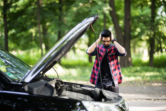 Closeup portrait, young indian man having trouble with his broken auto, opening hood trying to fix engine, isolated green trees outside background.