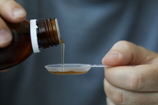Close-up of male hands holding holding bottle of cough expectorant syrup and pouring it with measuring spoon. Ill man taking medicine. Healthcare and treatment concept. Blurred background