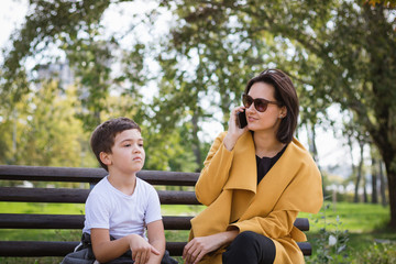 Mother talking on the phone while sitting with her son on park bench.