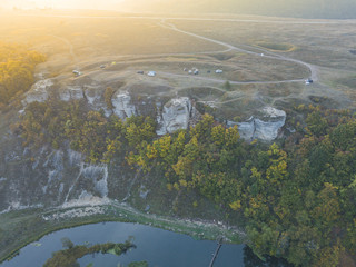 The turn of the river near rocks and cliffs on riverside covered by green forest and meadows and touristic camp with tents, cars and domes. Aerial view, drone shot. Russia, Vorgol river.