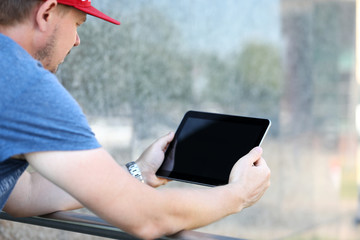 Focus on male in casual t-shirt and cap standing, leaning on railing. Stylish guy looking at screen of modern gadget. Copy space on display. Blurred background