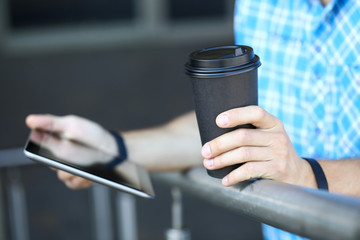 Focus on male hands holding paper cup of coffee and modern tablet. Stylish guy in casual t-shirt and fitness bracelet standing and leaning on railing. Blurred background