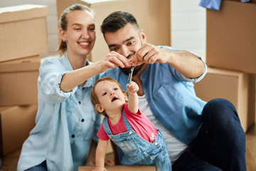 Fototapeta premium Family sit on floor near open box. Man and woman hold keys. Child want to take it. Background moving boxes
