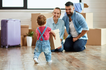 Kid wear denim overalls carries little moving box parents who sit on floor and smile