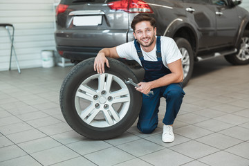 Naklejka na ściany i meble young handsome mechanic in uniform working in car service department fixing flat tire looks pleased