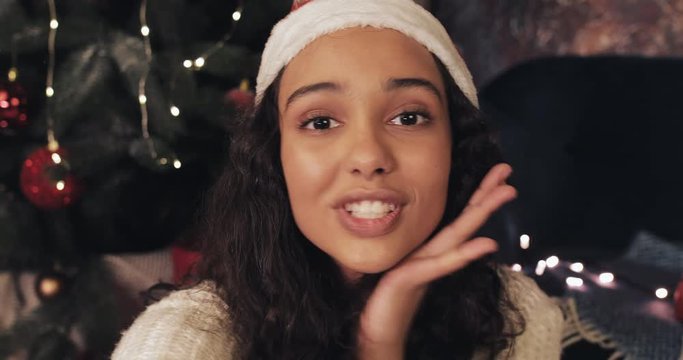 Attractive Young Smiling Woman in Santas Hat Making Selfie on her Smartphone Sitting at Home near Christmas Tree Background . New Year and Christmas Celebration Concept.