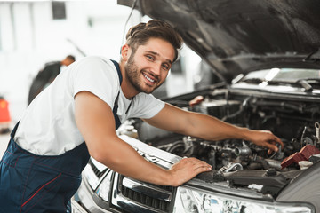 young smiling handsome mechanic in uniform fixing motor in car bonnet working in service center