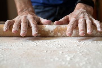Man's hands are rolling out dough in flour with rolling pin in her home kitchen. Homemade noodle or pasta production by father. Closeup, selective focus