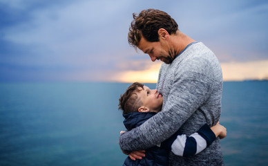 Father with small son on a walk outdoors standing on beach at dusk. - obrazy, fototapety, plakaty