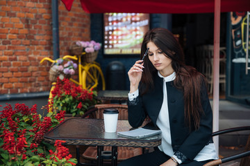 Young beautiful brunette girl, office clothing style, sitting on the street at a table in a cafe, street model posing style, makes notes in a notebook, talking on the phone