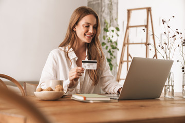 Woman sit indoors in office using laptop computer holding credit card.