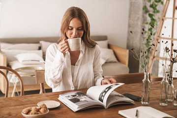 Woman sit indoors in office reading magazine drinking coffee.