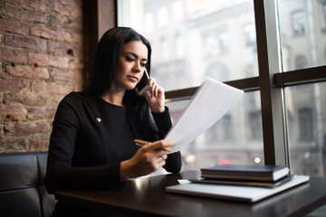 Fototapeta premium Confident woman skilled marketing coordinator reading paper documents and talking with client via mobile phone, sitting in restaurant interior. Female project manager exploring summary during work day