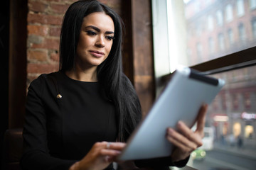 Woman candidate with good mood online booking via portable touch pad, sitting in restaurant during recreation time.Fashionable female blogger reading article via digital tablet, resting in coffee shop