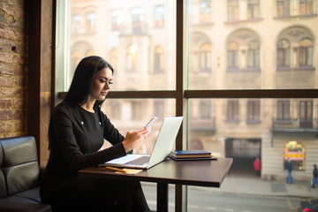 Young gorgeous fashionable woman reading pleasant text message on mobile phone during freelance work on pc laptop computer sitting in restaurant near window with copy space during recreation time