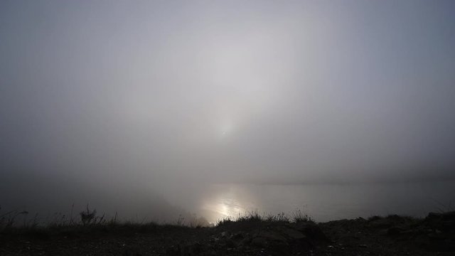 Sun breaks through the fast-moving fog on the river. Bright glare of light on the water. Steep coast. Gloomily. Timelapse.