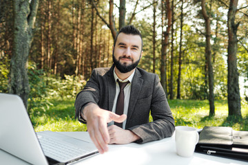 Young attractive business man in suit and tie sits at desk and works on computer outdoors. Gesture of reaching out and shaking hands.