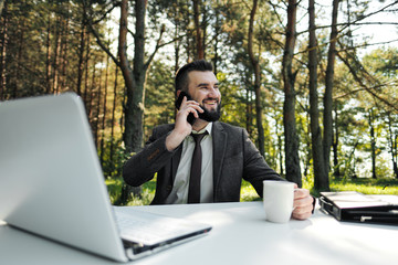 Young attractive business man in suit and tie sits at desk and works on computer outdoors. Drink coffee from white cup and talks phone. Green trees, nature and park on background.