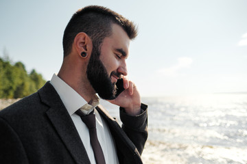 Young attractive male businessman in a brown suit with a briefcase stands on the seashore. Unfastens shirt, loosens tie and raises hands to sea.