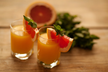 2 clear glasses with orange juice on the edge of the glass a piece of grapefruit and a mint leaf on a wooden background