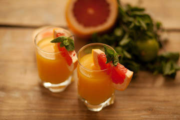 2 clear glasses with orange juice on the edge of the glass a piece of grapefruit and a mint leaf on a wooden background